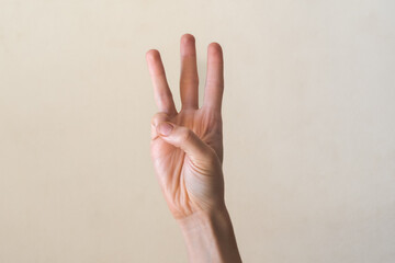 A hand with a light skin tone is raised, showing the number three with fingers extended. The background is a neutral color, emphasizing the gesture.