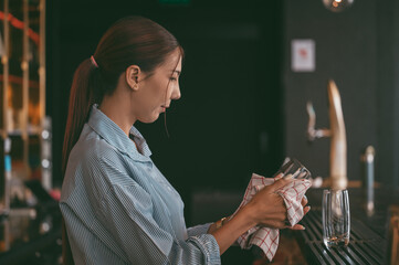 Young woman bartender working behind the counter, carefully cleaning a glass with a towel in a modern bar. restaurant industry concept.