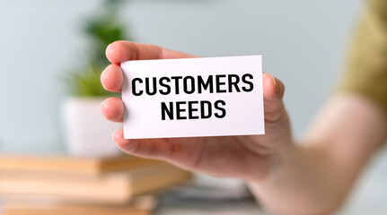 A hand holding a white card that says Customers Needs