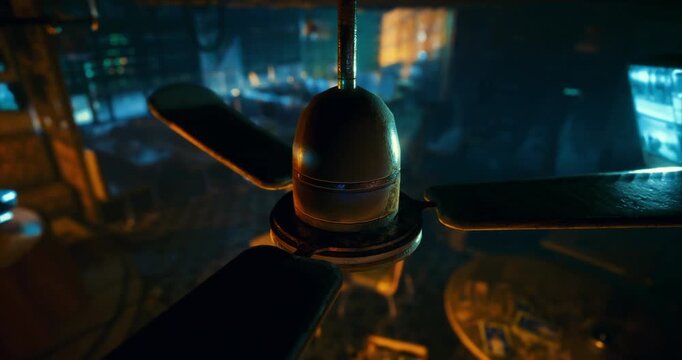 A close up view of a ceiling fan reveals its metallic texture and blades, set against a backdrop of softly illuminated furniture in a cozy, dark room.