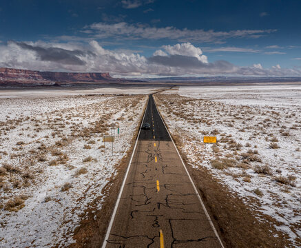 Aerial view of a cracked road cuts through the snowy desert landscape, leading towards distant mesas under a vast, cloudy sky, Sedona, Arizona, United States.
