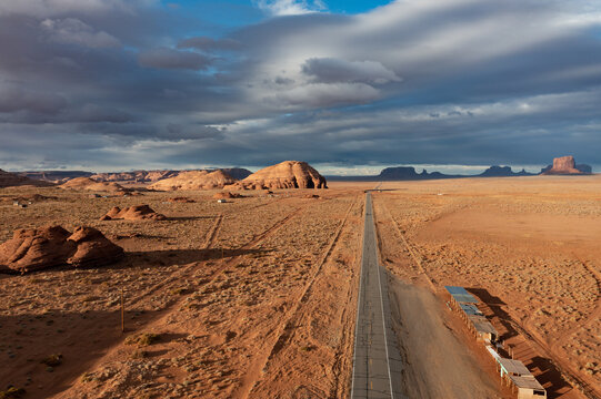Aerial view of a long, gray road stretching through the vast, arid landscape towards the iconic red rock formations under a dramatic sky, Sedona, Arizona, United States.