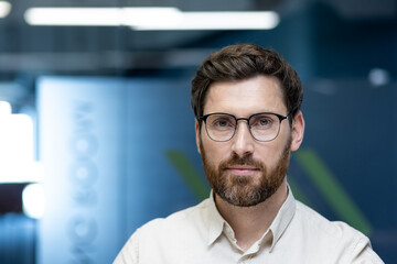 Close-up portrait of a young man in glasses and a beard sitting in an office, looking seriously and confidently at the camera