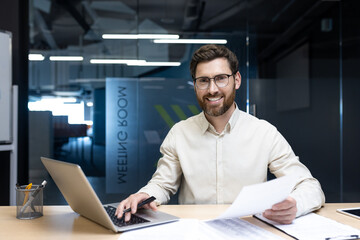 Smiling young businessman in glasses and shirt sitting at a desk in the office, working on a laptop, holding documents in his hand and looking at the camera