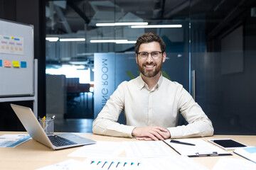 Portrait of a young man sitting at an office desk with a laptop and documents, looking and smiling at the camera.