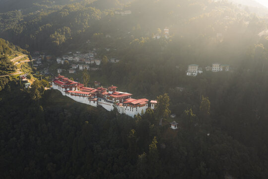 Aerial view of majestic Trongsa Dzong perched atop a hill, bathed in golden light against the backdrop of lush, dark green forests, Trongsa, Trongsa, Bhutan.