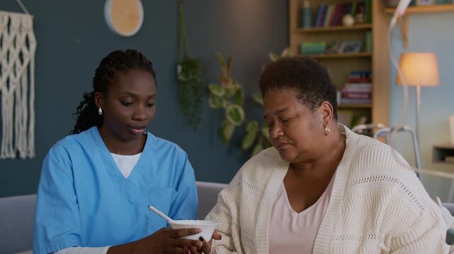 Medium shot of young female black caregiver feeding elderly patient with porridge during breakfast at home