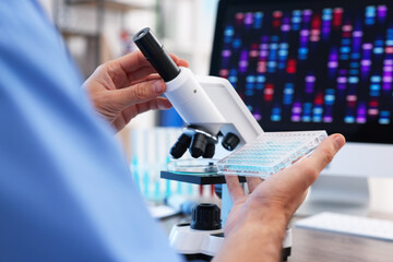 Research in genetics. Scientist working with microscope and microplate at table in laboratory, closeup