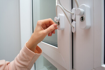 Woman's hand turning white window lock with safety cable. Modern home security latch for child protection and burglary prevention.