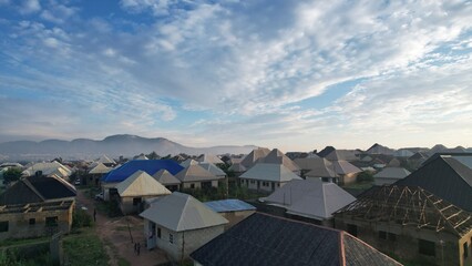 Aerial view of rooftops and homes nestled under a vast, cloudy sky, with distant hills adding depth to the scene, Abuja, Nigeria.