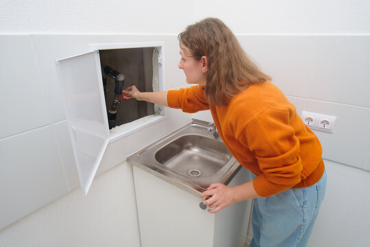 Woman plumber turning off water valve under sink. Female hand adjusting pipe in utility closet. Home repair emergency concept.