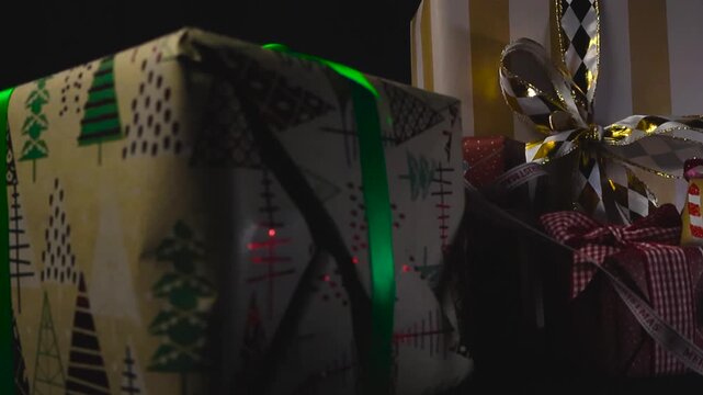 Closeup view sweeps along row of wrapped Christmas presents adorned with ribbons and bows against black background under studio lights. Glossy rosettes fall in slow motion, bouncing off the gift boxes