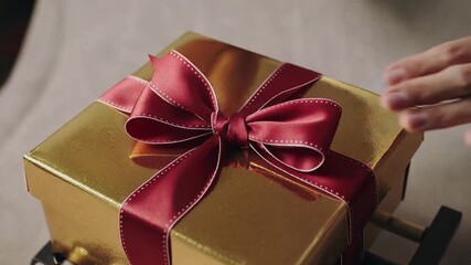 Hands hovering over gold gift box pinching red ribbon knot on wrapping table, forming bow - Powered by Adobe