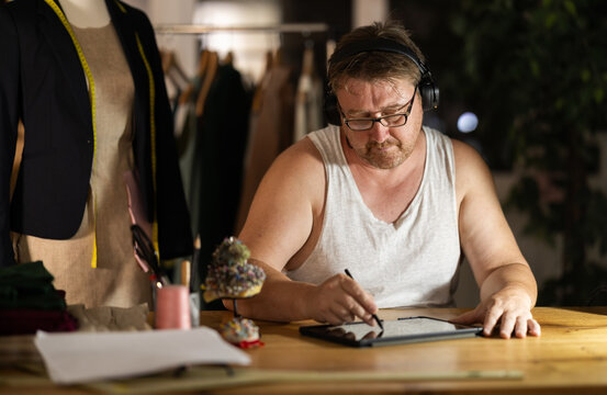 Middle-aged man wearing earphones drawing on tablet in sewing workshop at night