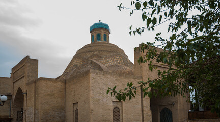 Fototapeta premium Historic domed building with a distinctive turquoise cupola and intricate brickwork, part of the architectural ensemble in the old town of Bukhara, Uzbekistan, framed by leafy tree branches in summer.