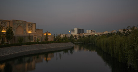 Obraz premium Silk Road complex in Samarkand at twilight with illuminated traditional architecture, willow trees along a canal, modern buildings in the distance, and full moon rising above the skyline of Uzbekistan