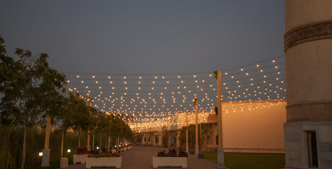 Pathway in the Silk Road complex of Samarkand at dusk with decorative street lamps and trees, illuminated by hanging string lights above, with flower boxes and historic walls in the background. © eskstock
