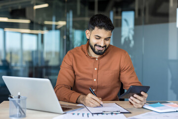Indian businessman smiling at desk while reviewing paperwork with laptop and calculator, managing budgeting, taxes and finances in a modern office environment