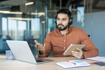 Man in headphones participating in an online video call meeting, talking and gesturing while looking at a laptop and holding a notebook in a modern office setup