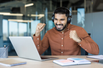 Obraz premium Young man wearing headphones happily celebrating business success or achieving a goal while looking at a laptop computer in a modern office workplace, fists pumping in excitement