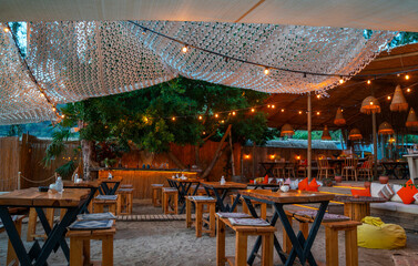 A cozy outdoor restaurant set on a sandy beach, adorned with wooden tables and benches, surrounded by lush greenery. Soft fairy lights and woven lanterns create a warm, inviting ambiance.