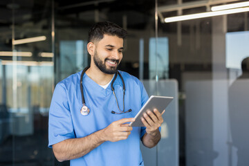 Young male doctor smiling, wearing scrubs and a stethoscope, interacting with a digital tablet and technology, managing healthcare data in a modern medical office setting