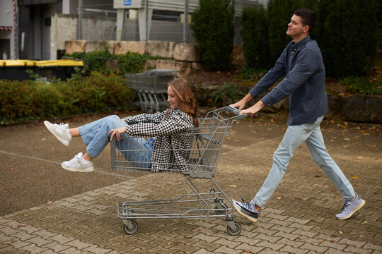 Playful Young Couple's Shopping Cart Adventure in Vibrant Park Setting. Joyful Outdoor Moment Captures Spontaneous Love and Seasonal Casual Style.