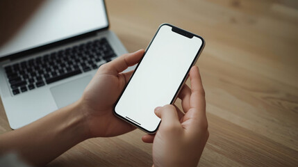 Mockup image of hands holding a black mobile phone with blank screen while using laptop computer in office