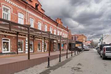 Historic brick buildings with modern awnings and string lights along a city street. Overcast sky and wet pavement suggest an approaching rain.