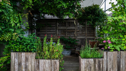 Cozy garden corner with lush greenery, wooden planters filled with herbs and flowers, a rustic wooden fence, and a shaded seating area, creating a natural and tranquil outdoor retreat.