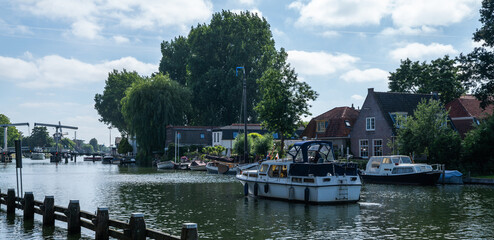 Obraz premium Boat navigating the canal in Weesp, Netherlands, passing moored vessels, brick houses, and green trees under a blue sky, with drawbridges and a distant windmill visible along the quiet waterway.