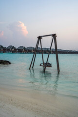 wooden swing suspended over shallow turquoise water near a sandy beach, with a row of over water bungalows on stilts in the background, creating a tropical paradise setting during a soft pastel sunset