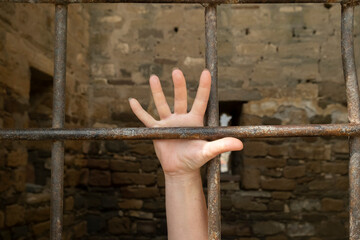 Hand reaching through rusty bars in an old stone prison cell