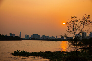 Peaceful sunset over a river with city skyline in the background, warm golden light reflecting on water, tranquil urban landscape blending nature and modern architecture.