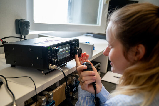 Young woman with ribbon in her hair operating a VHF/UHF amateur radio transceiver, holding a microphone and representing females and YLs in modern communication and STEM fields
