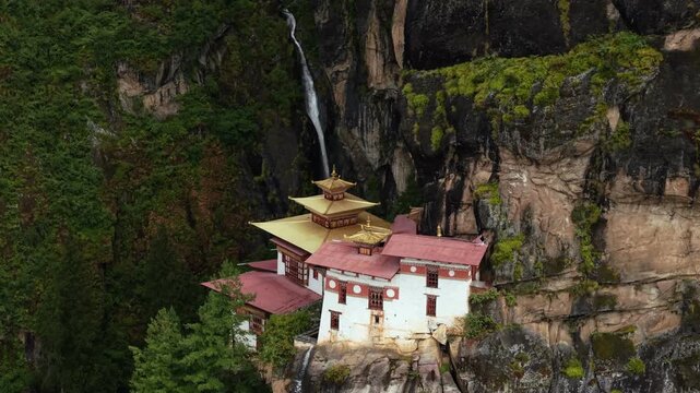 Aerial view of the Tigers Nest temple clings dramatically to a cliffside, contrasting with the lush greenery and cascading waterfalls, Paro, Bhutan.