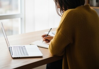 Woman writing in a notebook at a desk with a laptop. Student studying for an online course or working from home. Remote learning and freelance business concept