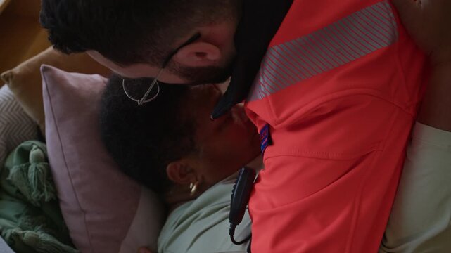 Close up shot of male paramedic in orange uniform radioing for assistance while placing elderly woman on pillow on couch