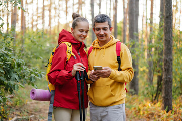 A couple stands on a wooded trail, smiling at the phone in their hands after finishing a hike in the forest during autumn