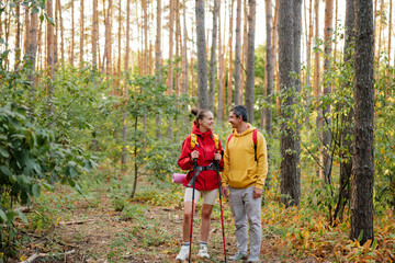 Two hikers smile at each other while exploring a vibrant forest filled with tall trees and sunlight