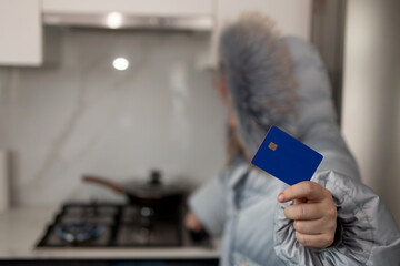 In a kitchen, a person in a puffy winter jacket holds a blue credit card. The gas stove is on in...