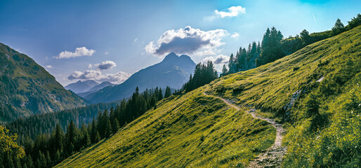 A hiking trail winds along the sunny slope of Mount Biberkopf with a view of the Rauhorn peaks....