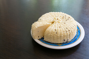 Close-up of round white homemade cheese wheel with textured surface and small slice cut out, placed on decorative blue-rimmed plate