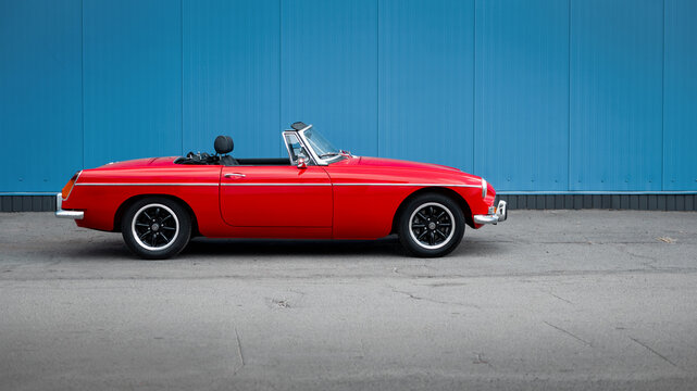 red MG MGB roadster on a street near blue wall. Side profile view of old 1960s open top british sports car. Kyiv, Ukraine - September 30 2024