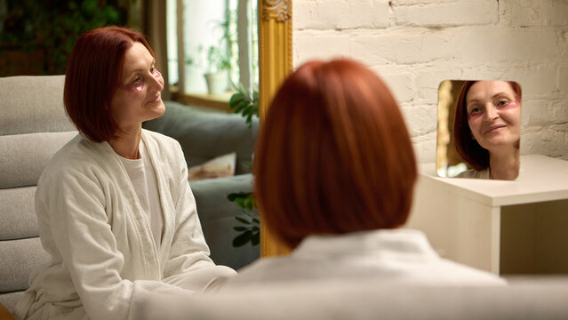 Grown woman with red hair using pink under-eye patches and smiling softly in natural light. Concept of confident aging, self-care rejuvenation, and emotional wellness in midlife beauty routine.
