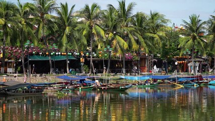 Riverside shops and cafes in Hoi An, Vietnam, feature colorful lanterns and traditional architecture, sheltered by tall palm trees, with tourist boats moored in the foreground.