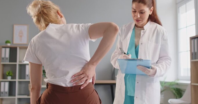 Young female doctor recording patients symptoms in medical card, listens and takes notes while patient explains her lower back pain discomfort. Healthcare. Musculoskeletal issues, physiotherapy