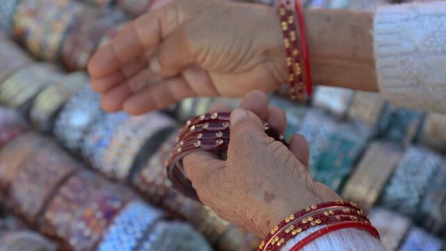 Close-up video shot of a bangle shopkeeper gently helping a village woman wear red bangles, surrounded by a vibrant array of colorful bangles at a lively rural Indian mela.