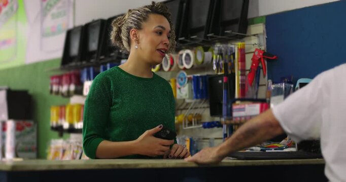 Female customer with curly hair using her smartphone to pay while interacting with an elderly male shopkeeper at the store counter during a transaction