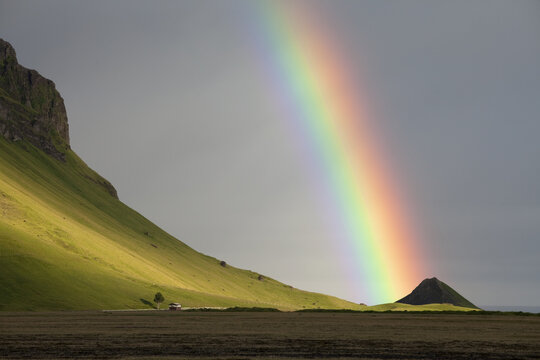 View of an arc of vibrant rainbow colors stretching across the sky, touching down on grassy slopes and a dark, rocky outcrop, Dyrholaey, Iceland.